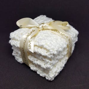 May include: A stack of four white crocheted washcloths tied with a cream-colored ribbon. The ribbon is printed with the words "Love by Lisa" and "Little Munchkins". The washcloths have a textured, handmade appearance.