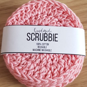 May include: A handmade, light pink scrubbie made of 100% cotton. The circular scrubbie is reusable and machine washable. The word "SCRUBBIE" is printed in bold, black letters on a white band wrapped around the scrubbie.