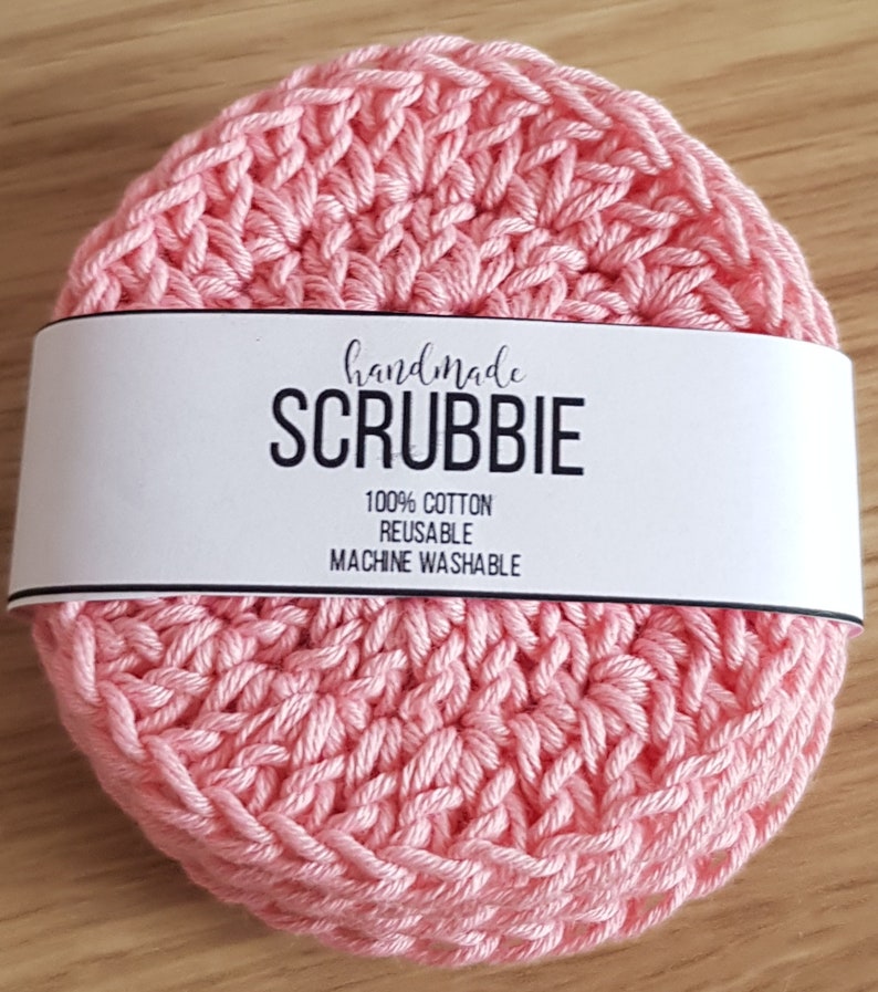May include: A handmade, light pink scrubbie made of 100% cotton. The circular scrubbie is reusable and machine washable. The word "SCRUBBIE" is printed in bold, black letters on a white band wrapped around the scrubbie.