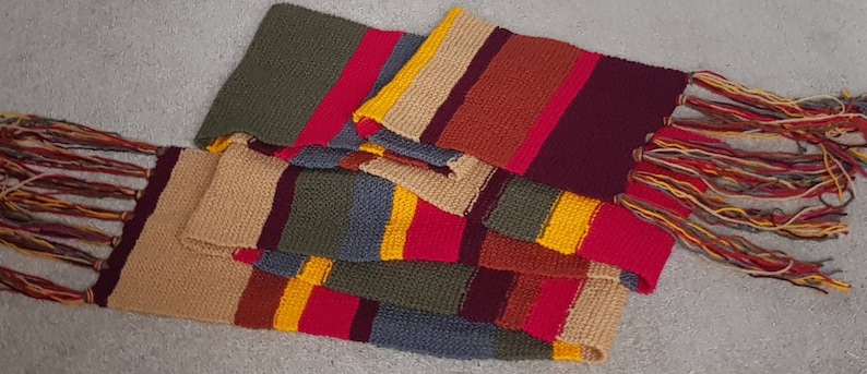 K&ouml;nnte beinhalten: Ein farbenfroher, rechteckiger Schal mit Streifenmuster. Der Schal hat Bl&ouml;cke in verschiedenen Farben, darunter Rot, Gelb, Gr&uuml;n, Beige und Burgunder. Die Enden des Schals haben lange, mehrfarbige Fransen.