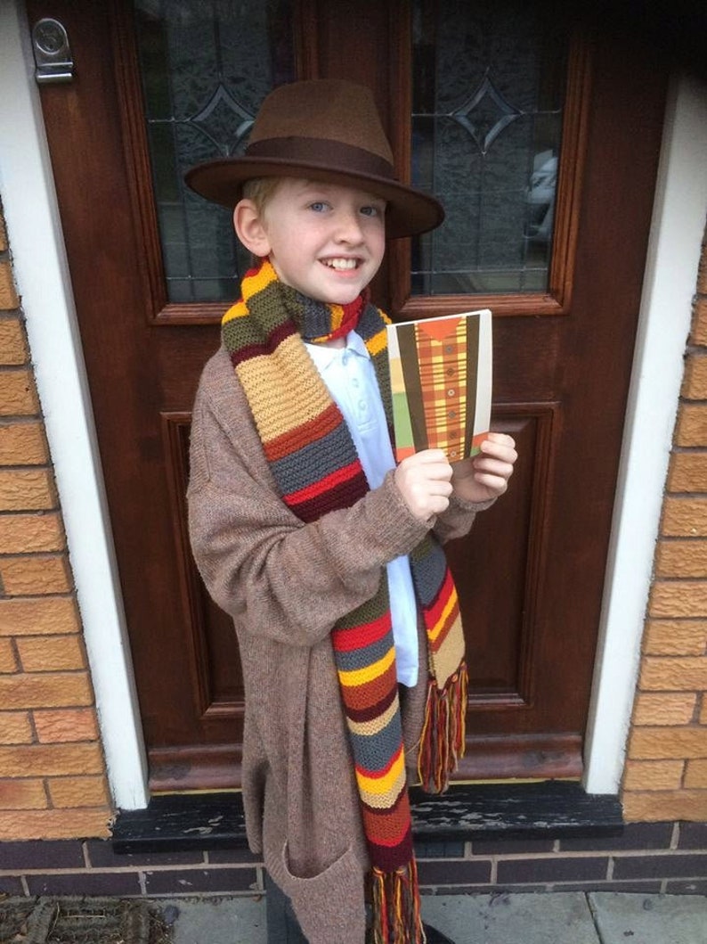 K&ouml;nnte beinhalten: Ein Kind tr&auml;gt einen braunen Fedora-Hut, einen langen, bunten Schal und eine braune Strickjacke. Der Schal hat Streifen in Rot, Gelb, Blau und Beige. Das Kind h&auml;lt ein Buch mit einem Karomuster.