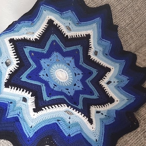 Può includere: Una coperta all'uncinetto fatta a mano a forma di stella, nei toni del blu, bianco e nero. La coperta presenta un design traforato dettagliato con un motivo circolare centrale e un bordo smerlato. La coperta è appoggiata su un divano grigio.