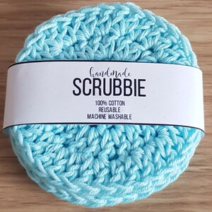 May include: A stack of handmade, light blue crocheted scrubbies. A white label wrapped around the scrubbies reads "handmade SCRUBBIE" with the text "100% COTTON REUSABLE MACHINE WASHABLE". The scrubbies are made of cotton.