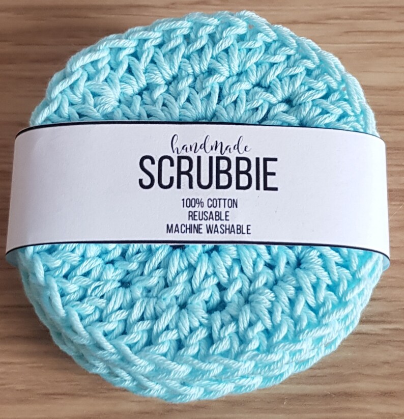 May include: A stack of handmade, light blue crocheted scrubbies. A white label wrapped around the scrubbies reads "handmade SCRUBBIE" with the text "100% COTTON REUSABLE MACHINE WASHABLE". The scrubbies are made of cotton.