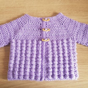 May include: A handmade, lavender crochet cardigan with long sleeves and wooden buttons. The lower section features a textured, bobble stitch design. A cozy, handcrafted baby or toddler sweater.