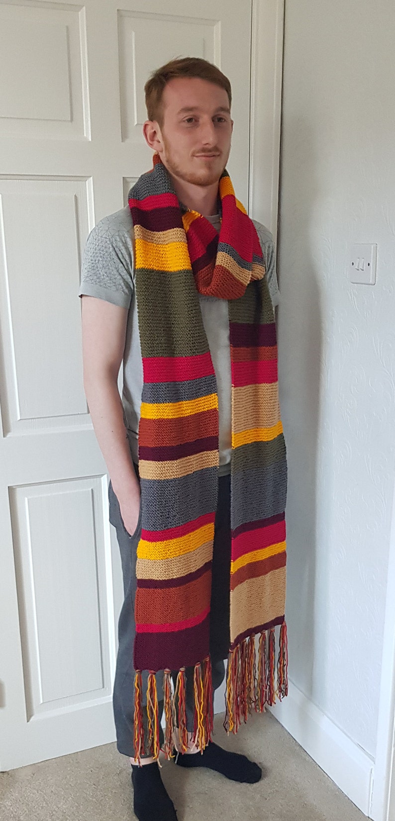 K&ouml;nnte beinhalten: Ein langer, gestreifter Schal in Rot, Gelb, Grau, Braun und Olivgr&uuml;n. Der Schal hat einen Fransenrand und wird um den Hals getragen. Ein markantes Accessoire f&uuml;r Fans einer bekannten Science-Fiction-Serie.