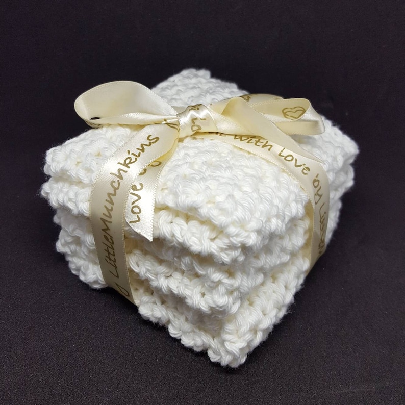 May include: A stack of white, crocheted washcloths tied with a cream-colored ribbon. The ribbon is printed with the words "Little Munchkins" and "Love by Lisa". The washcloths are square and have a textured appearance.
