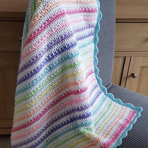 May include: A handmade, colorful crochet blanket draped over a gray chair. The blanket features horizontal stripes in pastel shades of pink, yellow, blue, and purple, with a scalloped light blue border. The blanket is a cozy, decorative item.