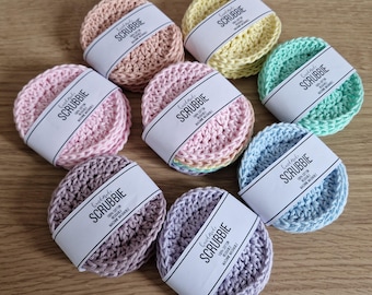 May include: A collection of eight handmade, circular scrubbies in various pastel colors, including pink, yellow, and blue. Each scrubbie is wrapped with a white label that reads "SCRUBBIE". The scrubbies are made of a knitted material.