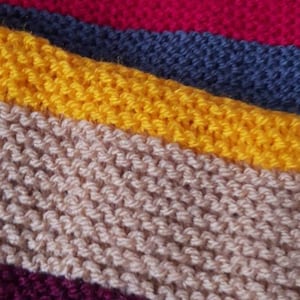 K&ouml;nnte beinhalten: Nahaufnahme eines Strickstoffs mit horizontalen Streifen in verschiedenen Farben. Die Farben umfassen Dunkelgr&uuml;n, Rot, Blau, Gelb, Beige, Lila und Rost. Die Textur des Stricks ist sichtbar.