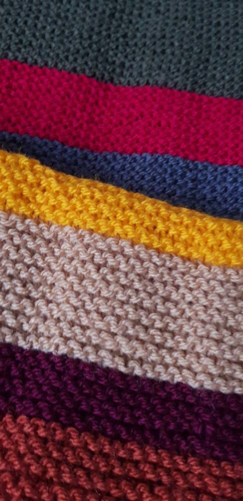 K&ouml;nnte beinhalten: Nahaufnahme eines Strickstoffs mit horizontalen Streifen in verschiedenen Farben. Die Farben umfassen Dunkelgr&uuml;n, Rot, Blau, Gelb, Beige, Lila und Rost. Die Textur des Stricks ist sichtbar.