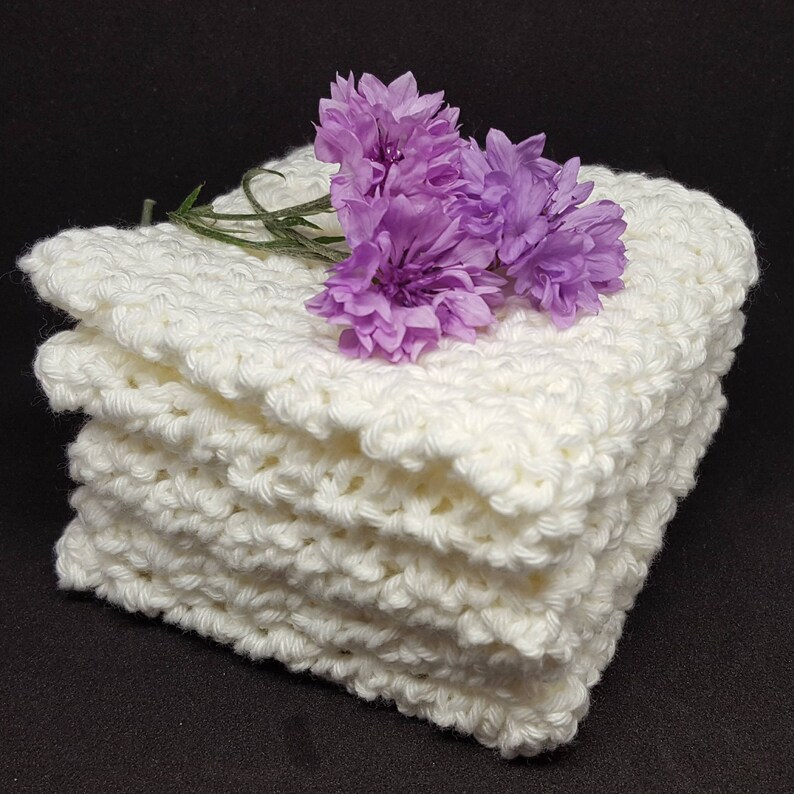 May include: A stack of four off-white, crocheted washcloths. The washcloths are made of a textured yarn and are stacked neatly. A sprig of purple flowers rests on top of the stack, adding a touch of color.