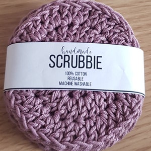 May include: A handmade, round, mauve-colored scrubbie made of 100% cotton. The scrubbie has a textured, woven appearance. A white label across the top reads "handmade SCRUBBIE" with details about the product's composition and care.