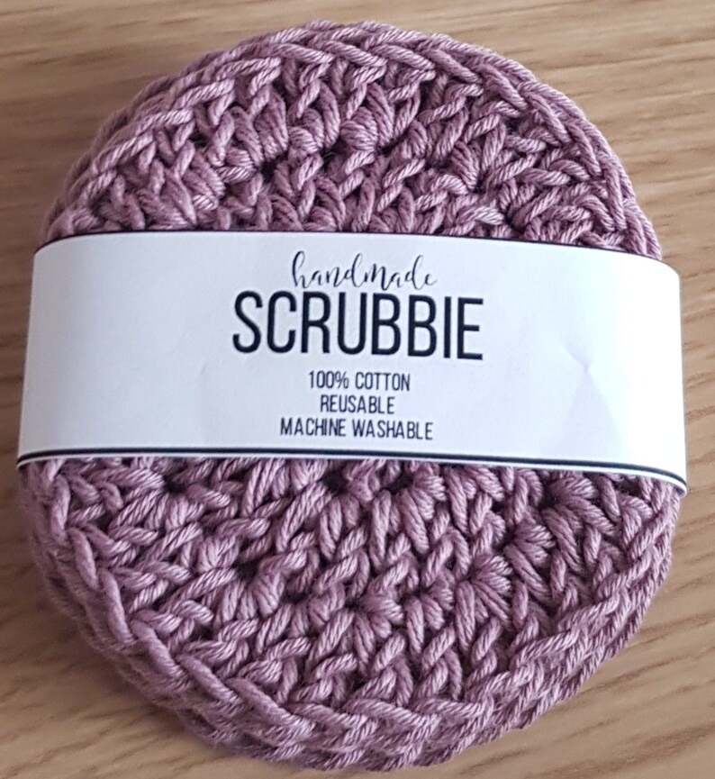 May include: A handmade, round, mauve-colored scrubbie made of 100% cotton. The scrubbie has a textured, woven appearance. A white label across the top reads "handmade SCRUBBIE" with details about the product's composition and care.