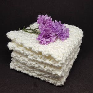 May include: A stack of four off-white, hand-crocheted washcloths. The washcloths have a textured, bumpy surface. A small sprig of purple flowers rests on top of the stack, adding a touch of color against the neutral background.