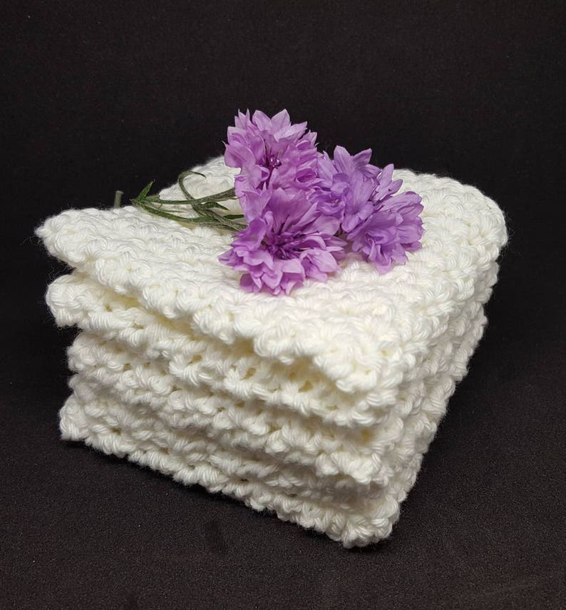 May include: A stack of four off-white, hand-crocheted washcloths. The washcloths have a textured, bumpy surface. A small sprig of purple flowers rests on top of the stack, adding a touch of color against the neutral background.