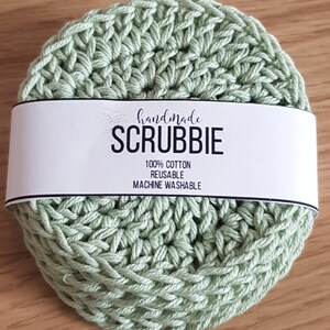 May include: A handmade, light green crocheted scrubbie with a white label wrapped around it. The label reads "handmade SCRUBBIE" with the text "100% COTTON REUSABLE MACHINE WASHABLE". The scrubbie is round and made of a textured, woven material.