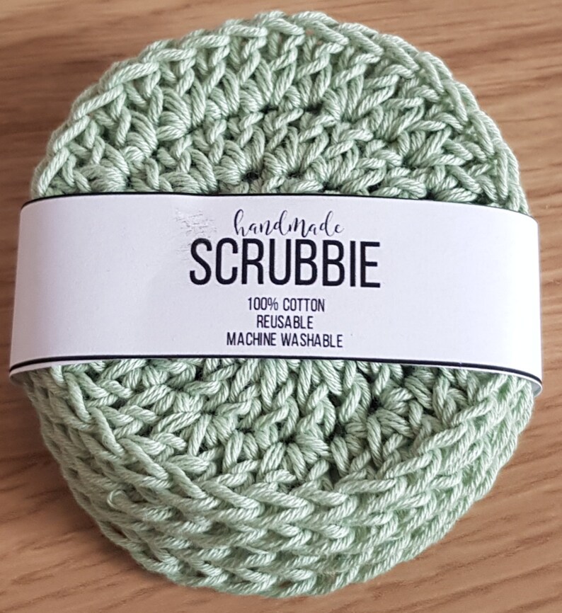 May include: A handmade, light green crocheted scrubbie with a white label wrapped around it. The label reads "handmade SCRUBBIE" with the text "100% COTTON REUSABLE MACHINE WASHABLE". The scrubbie is round and made of a textured, woven material.