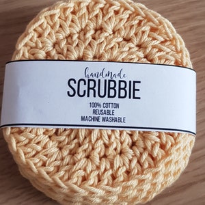 May include: A stack of handmade, light yellow crocheted scrubbies. Each round scrubbie is made of 100% cotton and is reusable and machine washable. The scrubbies have a white label that reads "handmade SCRUBBIE".
