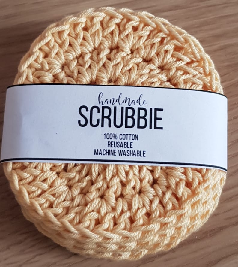 May include: A stack of handmade, light yellow crocheted scrubbies. Each round scrubbie is made of 100% cotton and is reusable and machine washable. The scrubbies have a white label that reads "handmade SCRUBBIE".