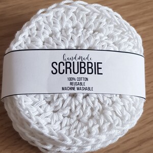 May include: A stack of white, handmade scrubbies made of 100% cotton. The reusable, machine washable scrubbies are round and crocheted. A white label across the stack reads "handmade SCRUBBIE".