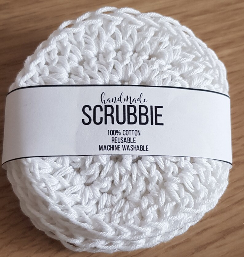 May include: A stack of white, handmade scrubbies made of 100% cotton. The reusable, machine washable scrubbies are round and crocheted. A white label across the stack reads "handmade SCRUBBIE".