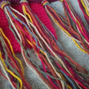 K&ouml;nnte beinhalten: Nahaufnahme eines geh&auml;kelten Schals in Burgunderrot und Fuchsia. Der Schal hat eine strukturierte Masche und farbenfrohe Fransen in Rot-, Gelb-, Blau- und Cremet&ouml;nen.