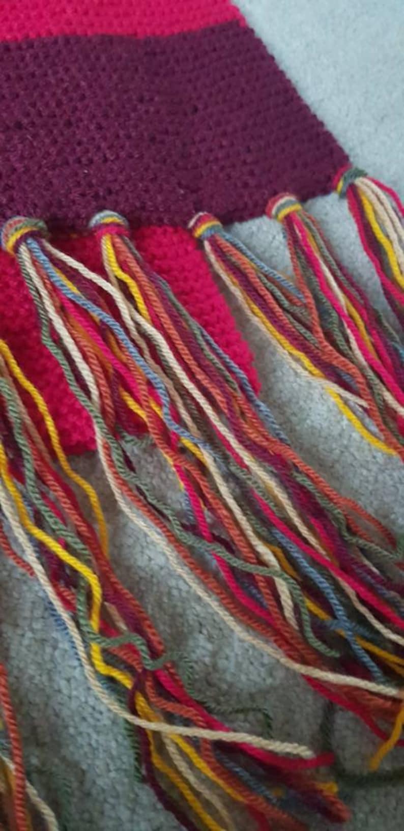 K&ouml;nnte beinhalten: Nahaufnahme eines geh&auml;kelten Schals in Burgunderrot und Fuchsia. Der Schal hat eine strukturierte Masche und farbenfrohe Fransen in Rot-, Gelb-, Blau- und Cremet&ouml;nen.