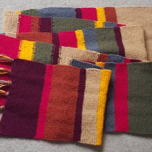 K&ouml;nnte beinhalten: Ein bunter, rechteckiger Schal mit Patchwork-Design. Der Schal hat verschiedene rechteckige Farbbl&ouml;cke in Rot, Gelb, Blau, Gr&uuml;n und Beige. Die Enden des Schals haben lange, mehrfarbige Fransen.