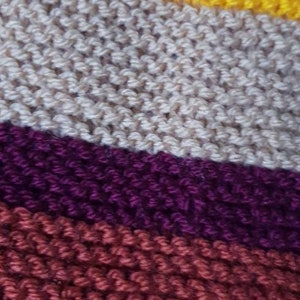 K&ouml;nnte beinhalten: Nahaufnahme eines Strickstoffs mit diagonalen Streifen in verschiedenen Farben. Die Farben umfassen Fuchsia, Blau, Gelb, Beige, Lila und Burgunder. Die Textur des Stricks ist sichtbar.