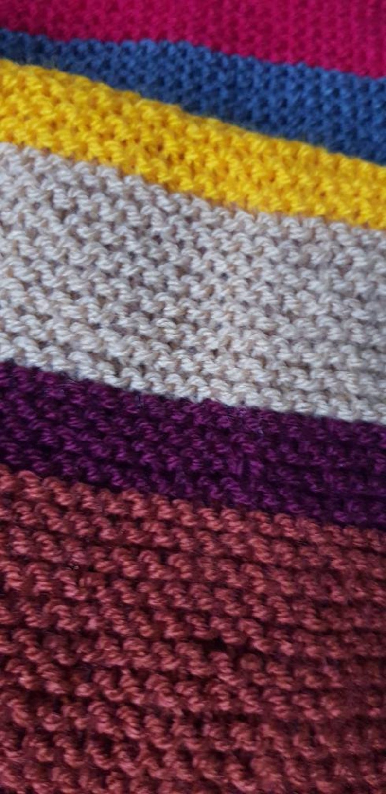 K&ouml;nnte beinhalten: Nahaufnahme eines Strickstoffs mit diagonalen Streifen in verschiedenen Farben. Die Farben umfassen Fuchsia, Blau, Gelb, Beige, Lila und Burgunder. Die Textur des Stricks ist sichtbar.