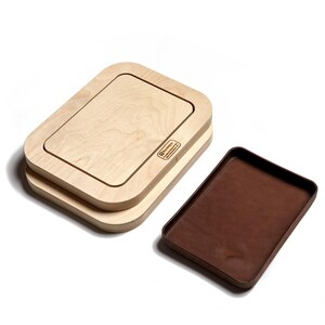 May include: A light-colored wooden press with rounded corners and a rectangular cutout, featuring the Buckleguy logo printed on the side. Beside it sits a separate, rectangular tray with a dark brown, smooth interior surface and raised edges.