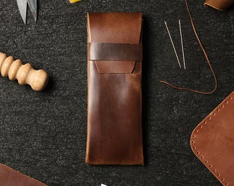 May include: A brown leather pencil case with a flap closure and a brown leather strap. The case is on a dark gray surface with other leather crafting supplies.
