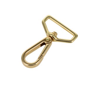 May include: A gold-toned metal swivel lobster clasp featuring a triangular-shaped loop at the top and a spring-loaded gate mechanism. The hardware has a smooth, polished finish and a sturdy construction designed for attaching straps or accessories.