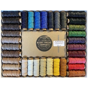 Puede incluir: Un conjunto de 36 carretes de cordón de poliéster encerado en varios colores, incluyendo marrón, beige, azul, verde, rojo y amarillo. Los carretes están dispuestos en una caja de madera con una etiqueta que dice "Maine Thread Company".