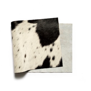 Puede incluir: Una pieza rectangular de piel de vaca con un patrón de manchas negras y blancas en la parte frontal. La textura del pelo es visible, mientras que el reverso del material muestra un acabado gris claro, mate y similar al ante.