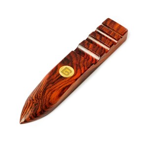 May include: A polished wooden tool with a dark reddish-brown grain pattern, featuring a pointed tip and a series of four parallel grooves along the top edge. A small, circular gold-colored emblem with a stylized letter G is embedded on the surface.