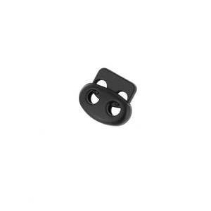 May include: A black cord lock with two oval-shaped holes and a rectangular section with two smaller holes. The cord lock is made of plastic and is designed to secure cords or drawstrings.