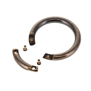 May include: A circular bronze-toned metal handle with a distressed finish, featuring a detachable curved segment and two small threaded screws for assembly. The hardware has a smooth, rounded profile and a weathered, antique-style metallic appearance.