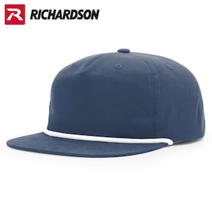 May include: A solid navy blue five-panel cap featuring a flat brim and a decorative white rope detail across the base of the crown. The Richardson brand logo is visible in the upper left corner, displaying a red R icon and the text RICHARDSON.