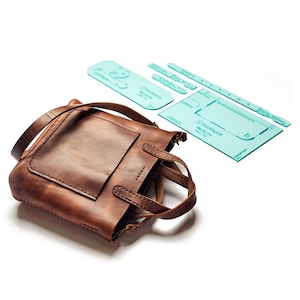 May include: A brown leather tote bag with a front pocket and handles. Beside it are several clear, light blue acrylic templates for bag construction. The templates are various shapes and sizes, including a rectangle and curved pieces.