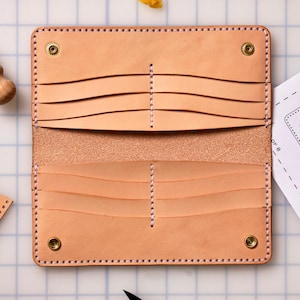 May include: Open, natural-toned leather wallet with multiple card slots and snap closures. The wallet is hand-stitched with a contrasting thread and features a textured interior. The design is minimalist and functional, showcasing the natural grain of the leather.