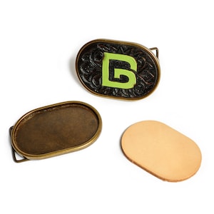 May include: A set of three oval-shaped belt buckles. One buckle is brown with a green letter "B" carved into it. The other two buckles are plain brass.