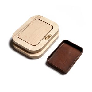 May include: A light-colored wooden rectangular tray with rounded corners and a matching wooden lid featuring the Buckleguy logo. Beside it sits a separate, smaller rectangular tray with a dark brown interior and a wooden exterior frame.