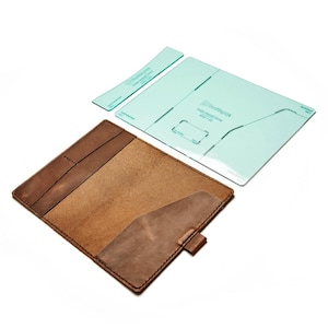 May include: A brown leather journal cover with a pocket and a snap closure. A light blue plastic template is next to the journal cover. The template has the text "Large Journal Cover" and other text.