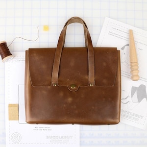 Könnte beinhalten: Eine braune Ledertasche mit zwei Tragegriffen und einem zentralen Messing-Druckknopfverschluss. Die Tasche hat einen rechteckigen Körper mit sichtbaren Nähten und Metallnieten. Sie wird zusammen mit einer Garnrolle und einem Holzpolierwerkzeug gezeigt.