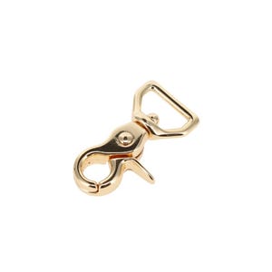 May include: A gold-toned metal swivel lobster clasp featuring a smooth, polished finish. The hardware includes a triangular loop at the top for strap attachment and a spring-loaded gate mechanism that opens inward to secure items firmly in place.