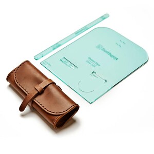 May include: A brown leather roll-up case with a strap closure, a clear ruler, and a teal-colored template with printed text. The items are arranged on a white background. The template has curved edges and cut-out shapes.