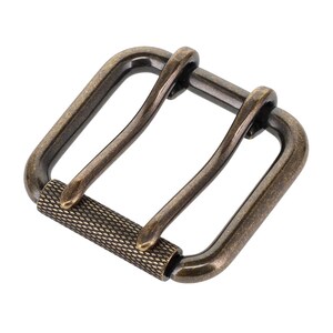 Può includere: Una fibbia per cintura quadrata con finitura in bronzo scuro. La fibbia presenta una cornice arrotondata e una barra centrale con un motivo testurizzato a tratteggio incrociato. Due ardiglioni curvi si estendono dalla barra superiore.