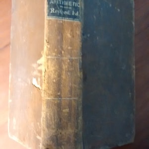 Puede incluir: Un libro antiguo con el título "DAVIES' UNIVERSITY ARITHMETIC Revised Ed." visible en el lomo. El libro tiene una cubierta marrón y una etiqueta azul con texto blanco, mostrando signos de edad y desgaste.